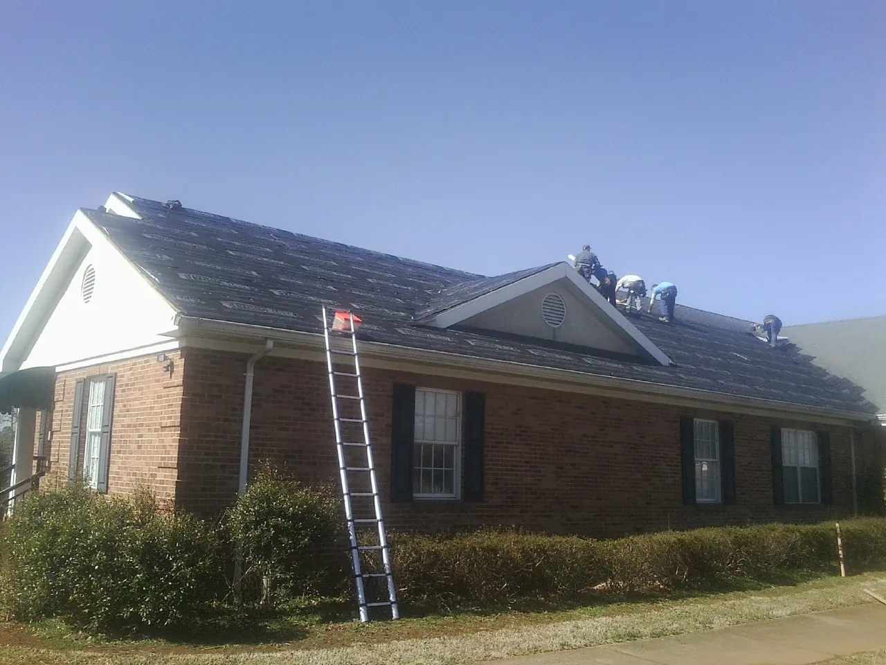 Crew tearing shingles off a brick ranch roof with a ladder set — Metal Roof Repair work by our crew in East Peoria