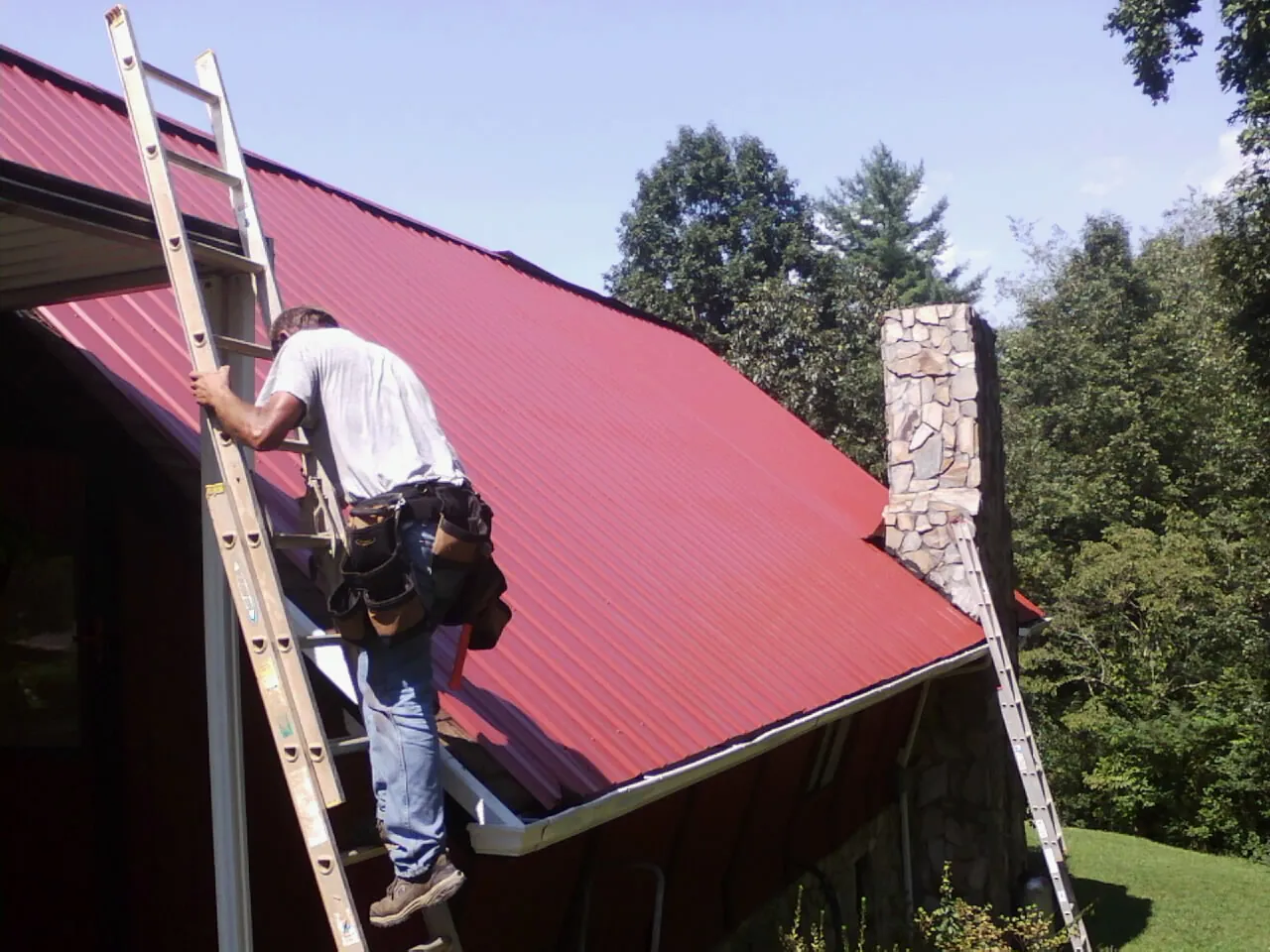 Roofer on a ladder beside a red metal roof and stone chimney — Soffit & Fascia Repair by Roofing Brothers in East Peoria